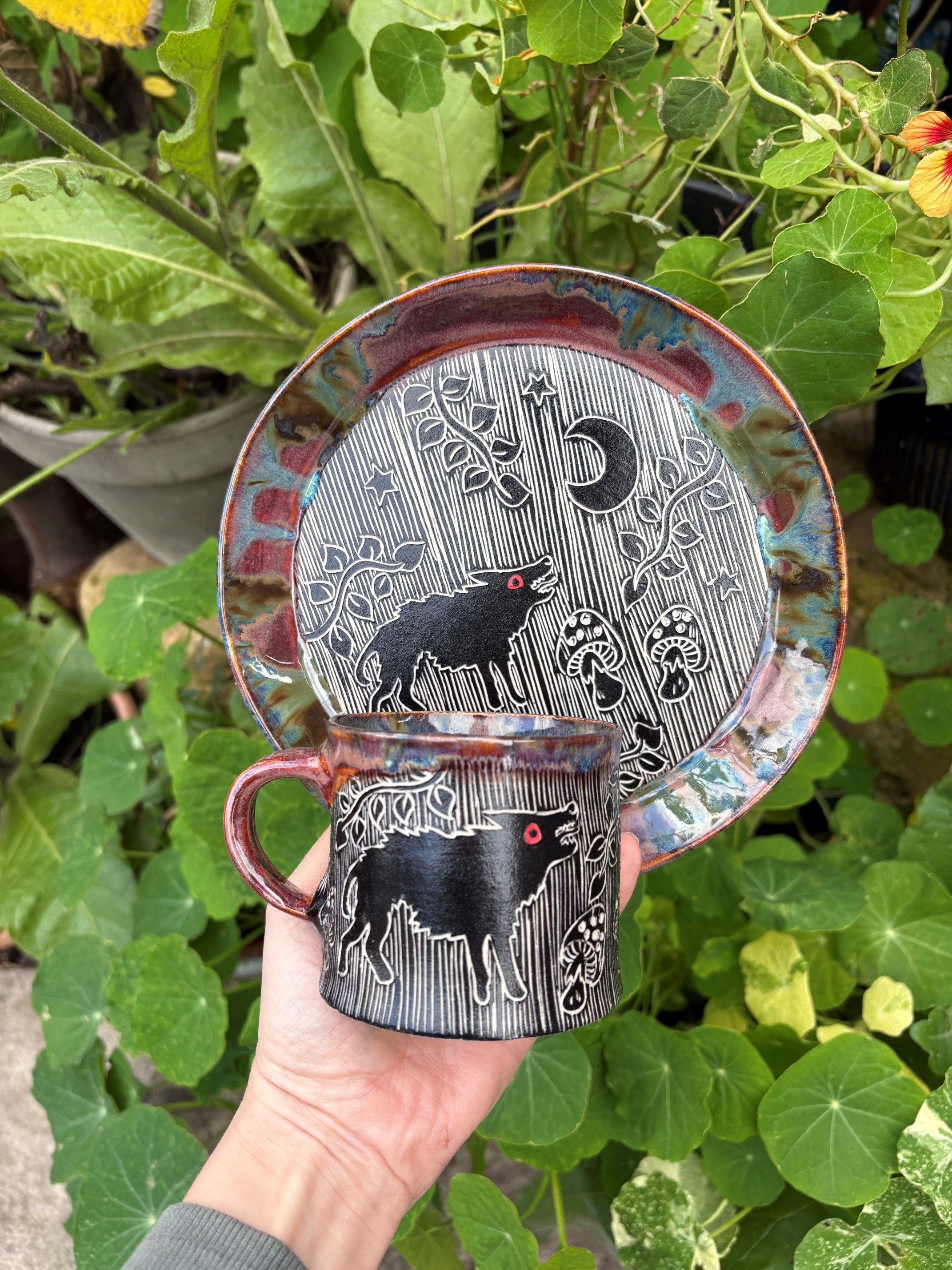 Image promoting sgraffito ceramics by Murtle and Thyme, with a cup and saucer showing Black Shuck, held in a hand against a leafy background. Picture by Charlie Spearing.