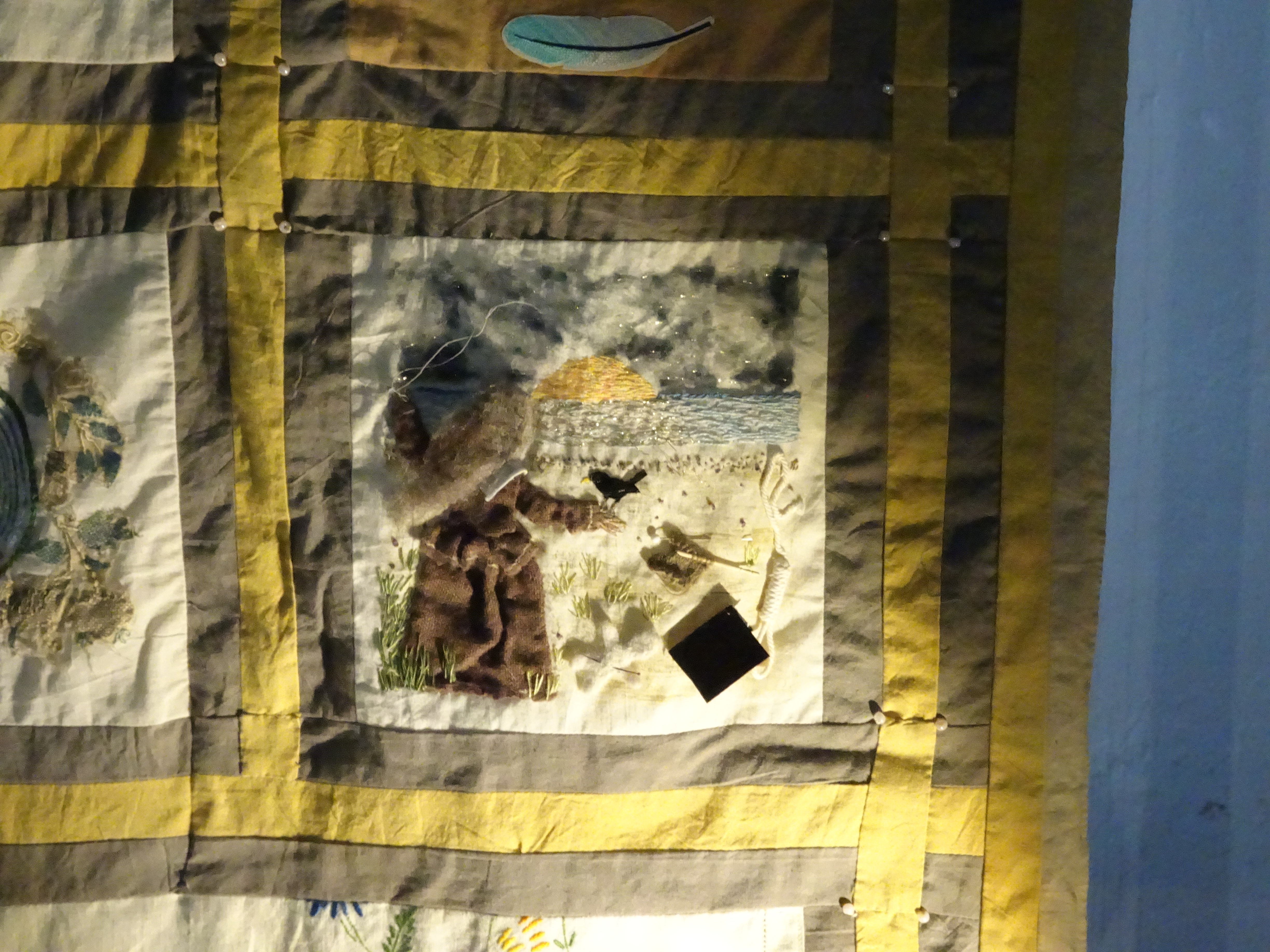 Quilt panel by Val Thomas showing a woman (Elizabeth Bradwell) with her back to us, looking across a beach at the rising sun, with a blackbird perched on her right hand, and to her right an intertwined noose and book and a piece of knitting with needles, above it.